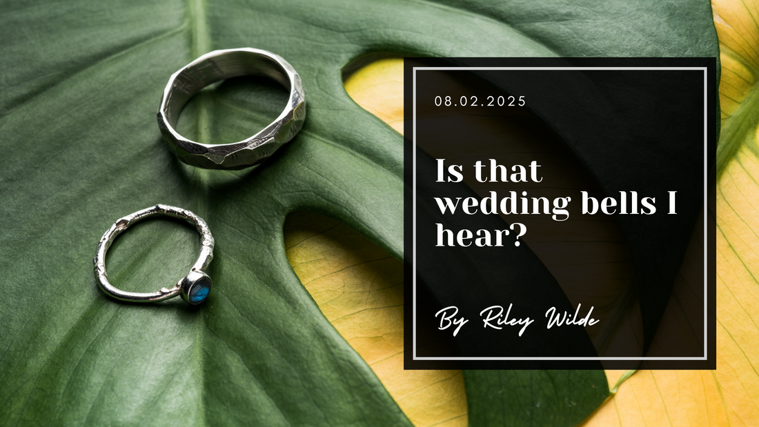 Two silver coloured rings rest on a dark and light leaf. A black box with white text holds the date 08.02.2025 and the title "Is that wedding bells I hear? By Riley Wilde".