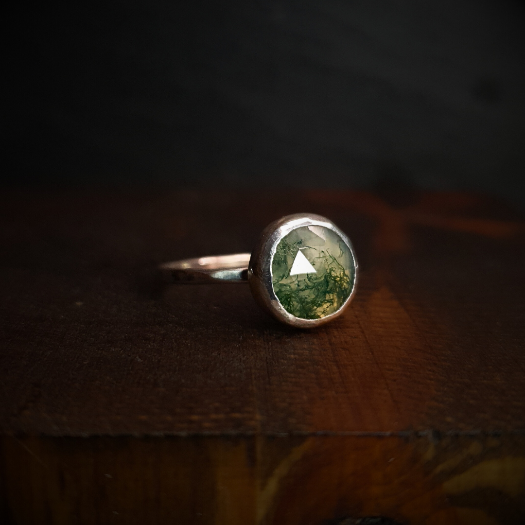 A moss agate ring sits against a dark background. It has a hammered silver band