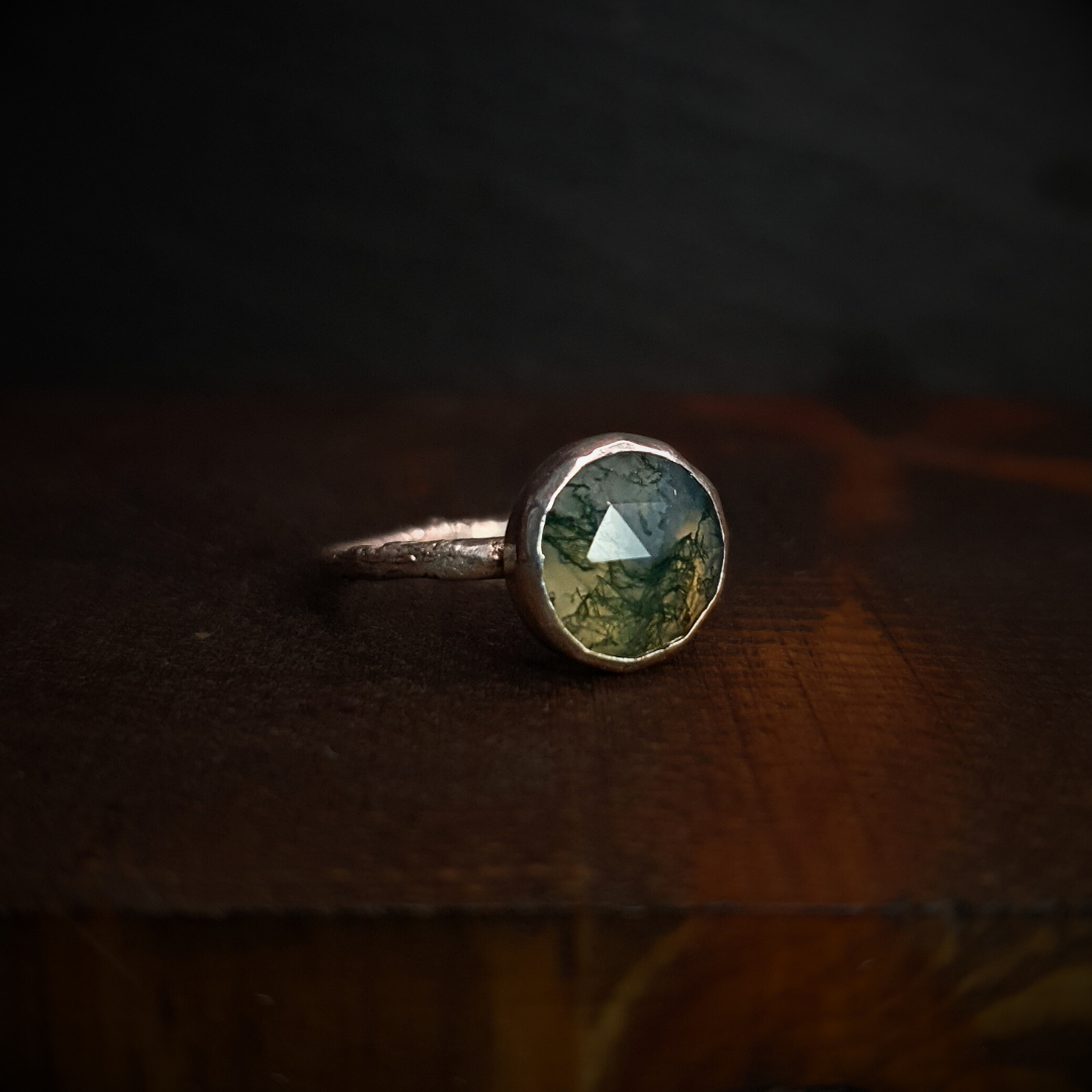 A moss agate ring sits against a dark background. It has a cast silver twig band.