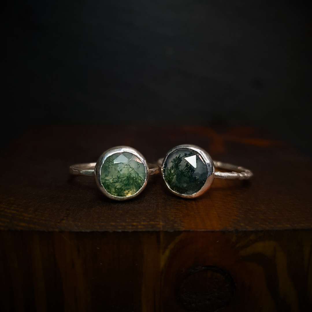 Two moss agate rings sit against a dark background. The one of the left has a hammered band and the one on the right has a twig band.