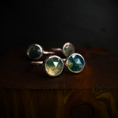 Four moss agate rings sit against a dark background. The two in the foreground have hammered bands and the two in the background have a twig band.