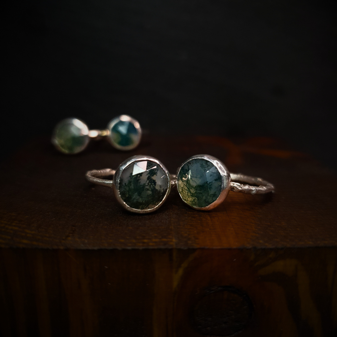 Four moss agate rings sit against a dark background. The two in the foreground have twig bands and the two in the background have a hammered band.